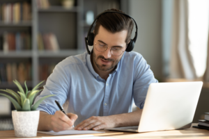 Focused 35s man sit at desk wear headphones watch webinar use laptop gain new knowledge writing notes.