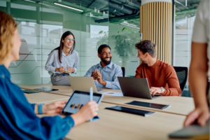 Diverse group of business professionals collaborating in a team meeting, sharing ideas, smiling, and using digital technology to support workplace communication and employee engagement.